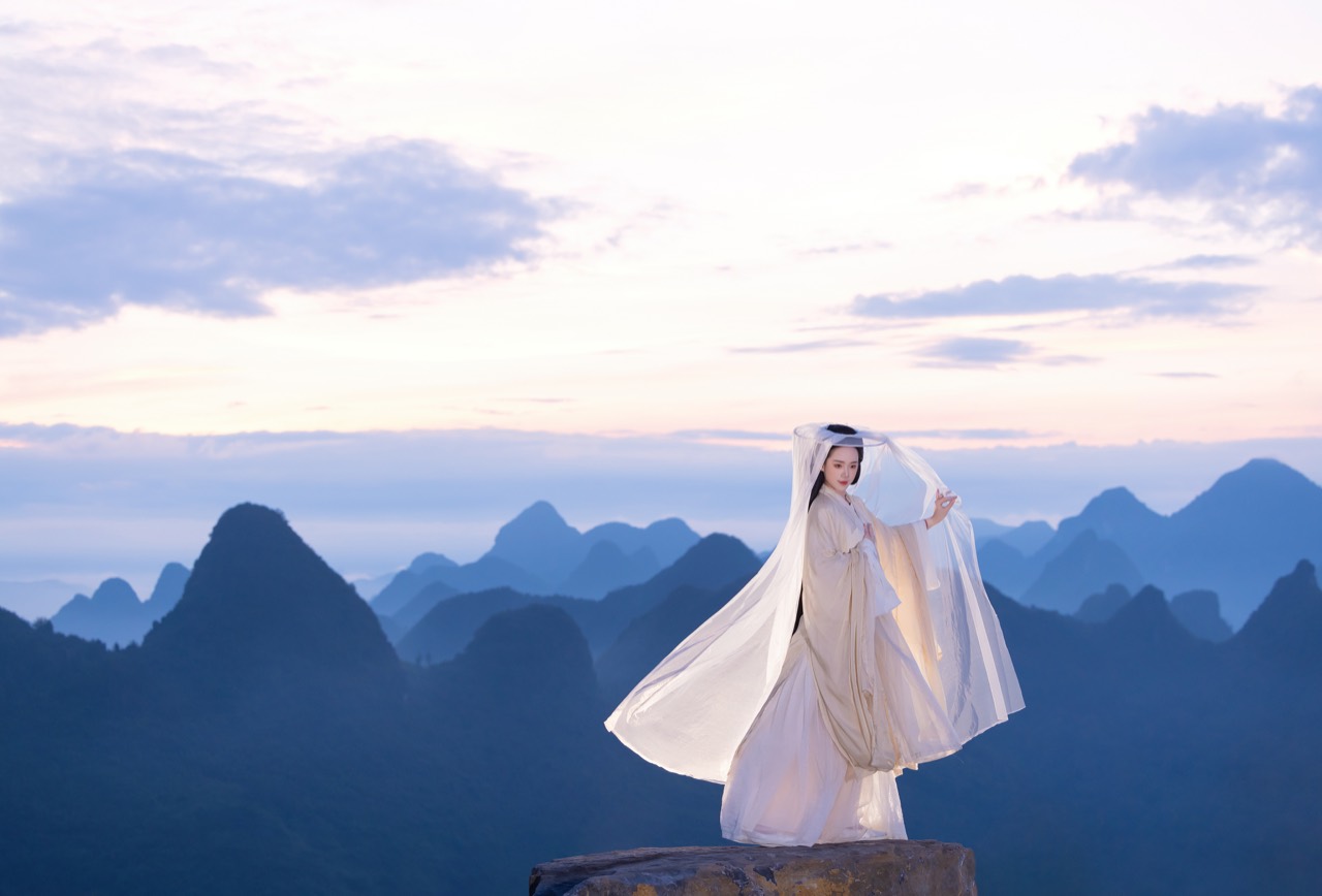 Chenshuo in flowing white clothing with mountains at sunset